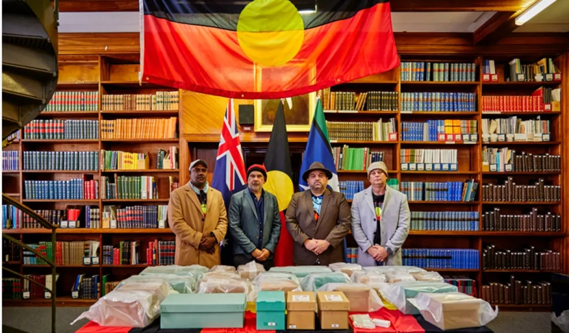 Four men stand behind a table under an Aboriginal flag