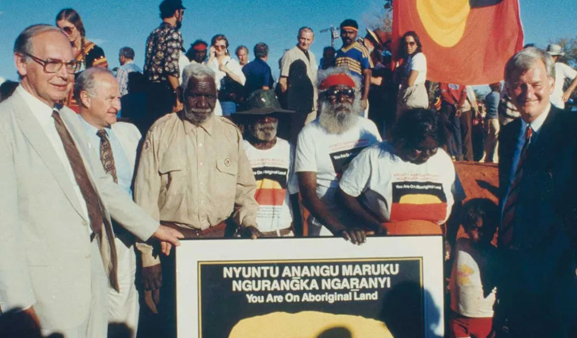 A group of people surround a poster plaque displaying the handing back of the land.