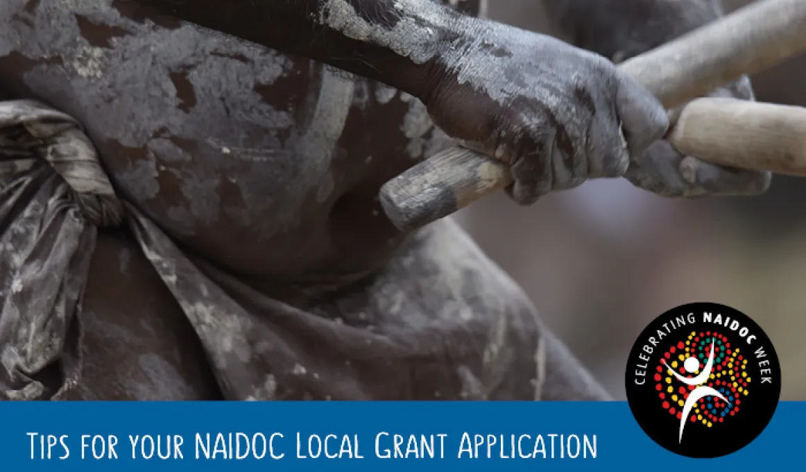 Closeup shot of hands holding sticks which are touching. The hands, arms and belly of an Indigenous person are painted in the traditional way.