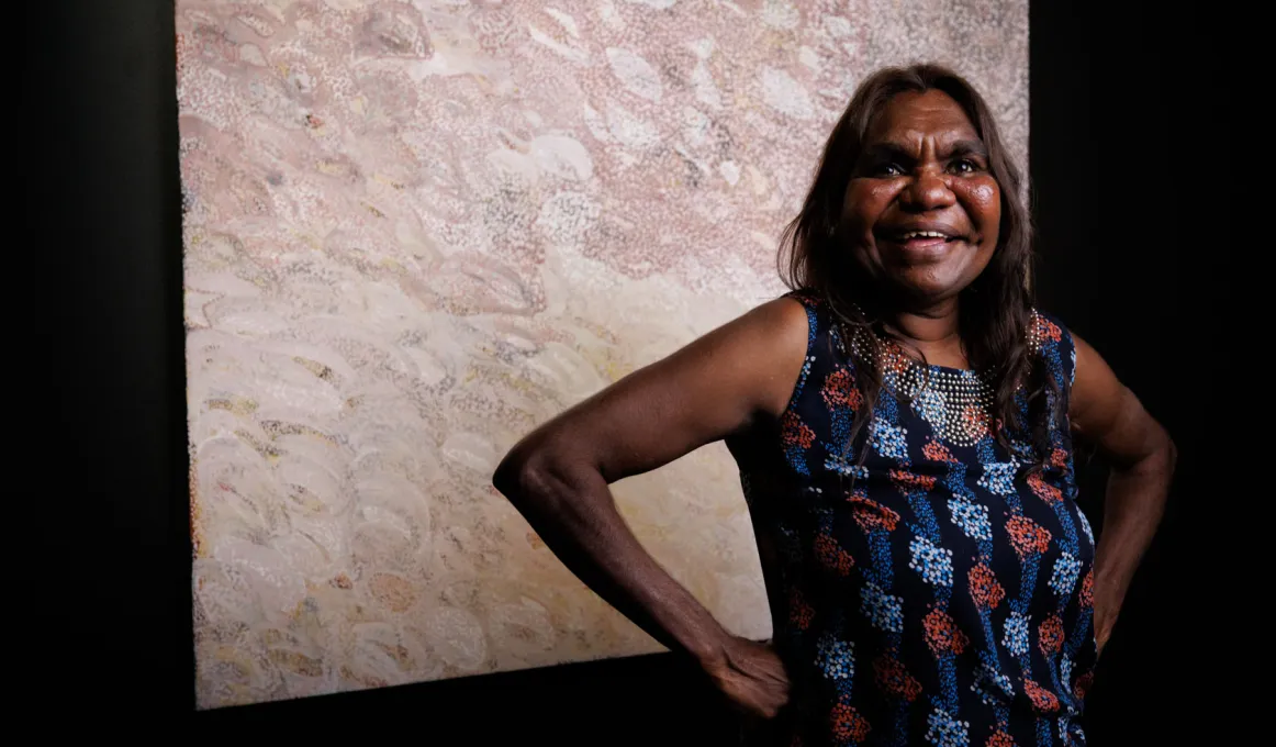 An Aboriginal woman stands with a broad smile and her hands on her hips. Her award winning piece is hanging on display behind her.