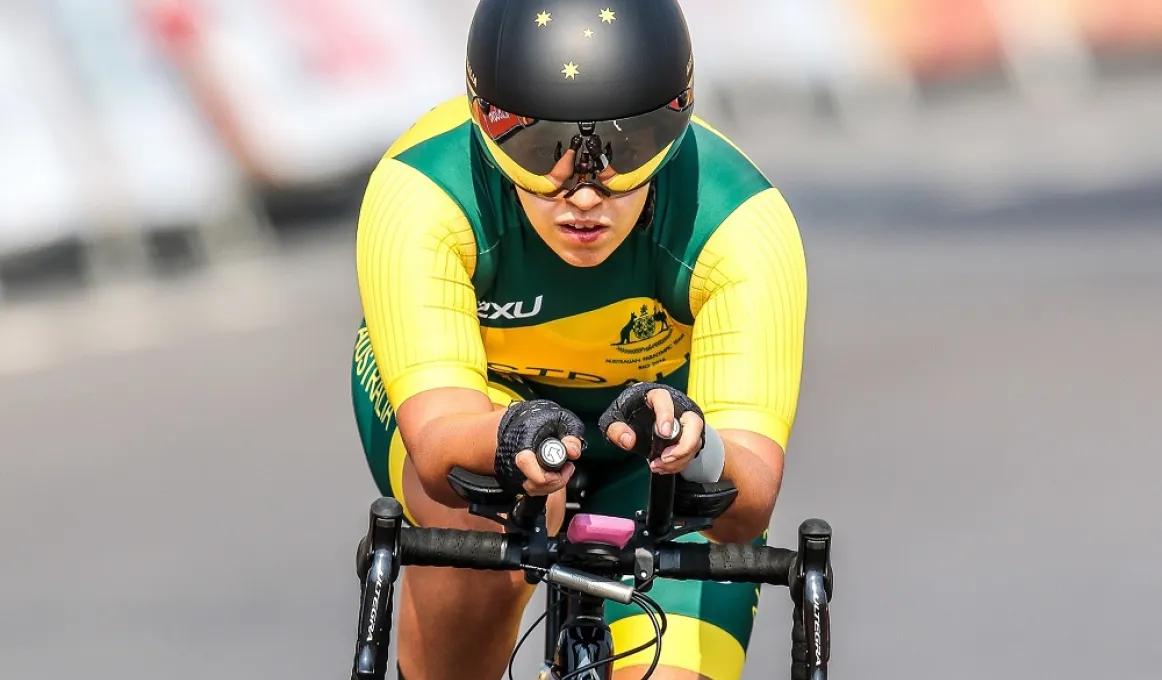 A woman dressed in green and yellow cycling wear rides a bike toward the camera. In the background is a dark road and signage.