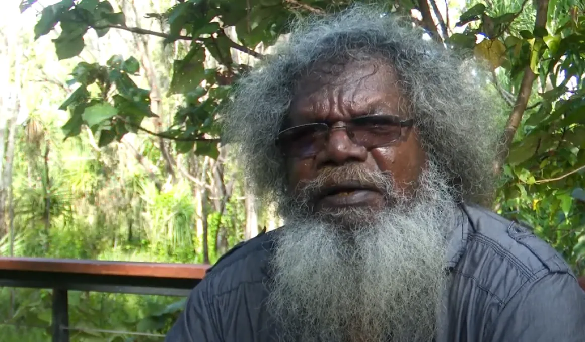 A man sits in front of a forest scene. He is wearing a grey-blue shirt and tinted glasses.