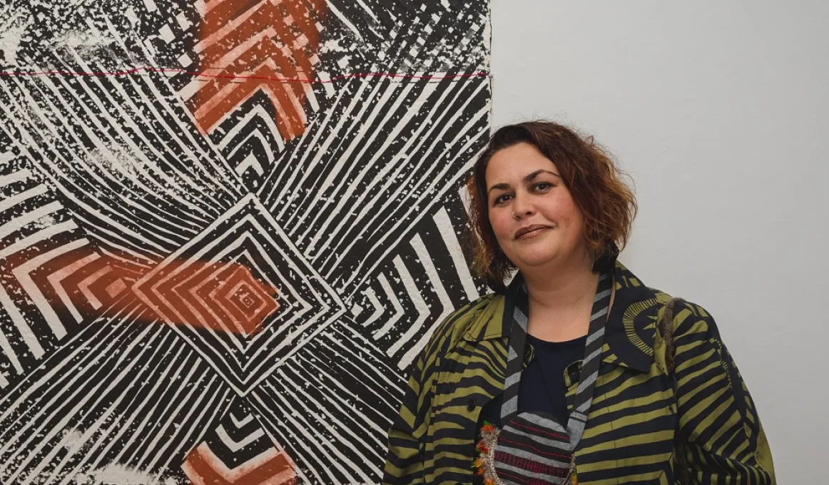A woman in a green and black striped coat stands next to a mixed media screen print on the wall. It is black, white and ochre in colour with lines and chevrons radiating from a centre made of concentric squares.