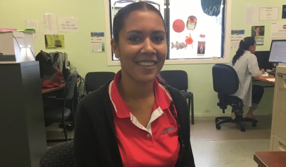 Young Aboriginal woman in red shirt sits in an office with equipment and her colleague in the background. The floor is grey and the walls green in colour.