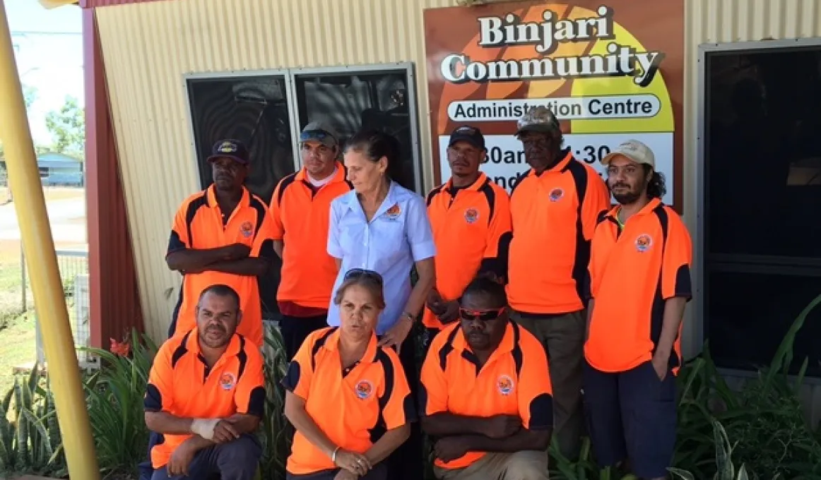 Two rows of nine Indigenous persons dressed in orange shirts (one in blue) stand and kneel in front of a yellow building with a sign on the wall which says, Binjari Community Administration Centre.