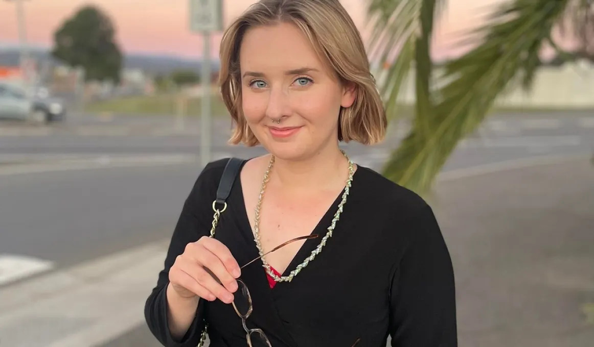 A young woman in a black top holds her glasses in front and smiles at the camera. In the background are tree branches, a car and some signposts.