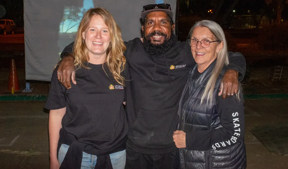 Two women and a man stand shoulder to shoulder. All are dressed in dark clothing. The background is dark except for a pale screen that displays a projected image.