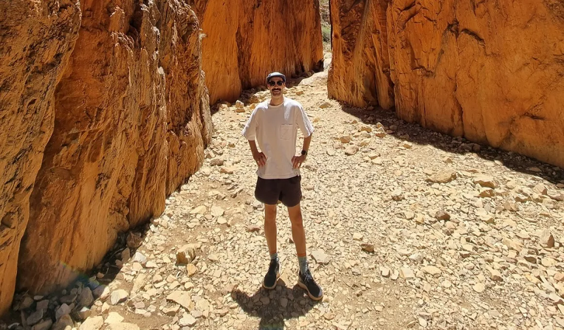 A man in light coloured shirt and dark shorts, wearing a cap and sunglasses, stands on rocky ground in a gorge with ochre coloured walls.