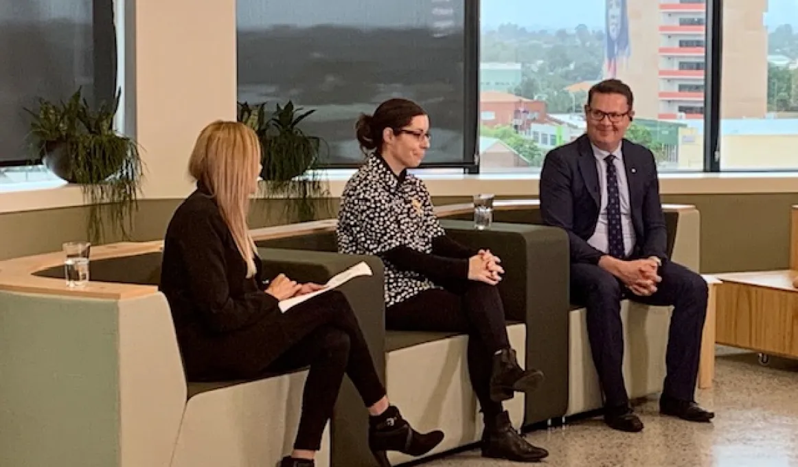 Two women and one man sit on couches in a room. In the background are windows and outside are buildings.