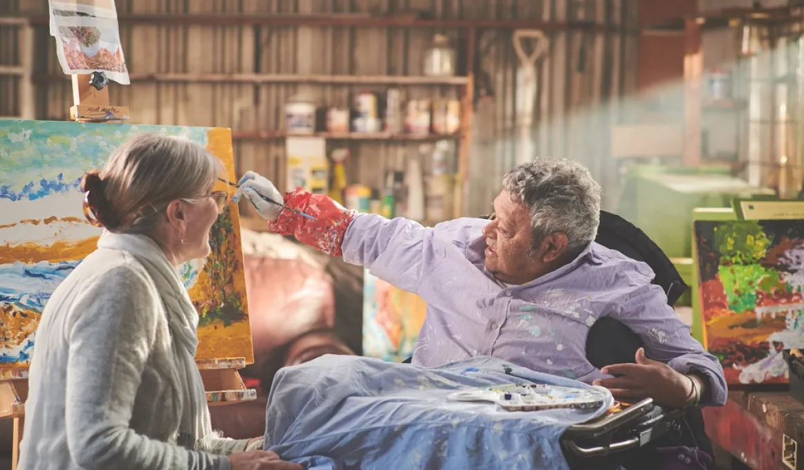 A woman and a man sit and talk to each other as the man paints a landscape picture. In the background is a wall with paints and other apparatus on shelving.