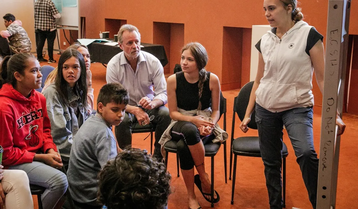 Aboriginal and non-Aboriginal adults and youth sit and stand in a large ochre coloured room, talking with each other. They are divided into two groups each with a whiteboard at which an individual stands and captures discussion points.