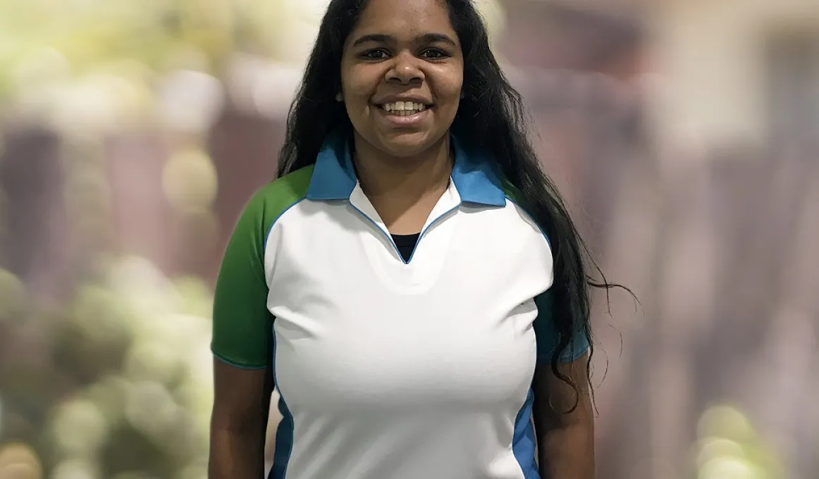 A young adult woman in a green, white and blue top smiles at the camera. She has long black hair. The background has been blurred.