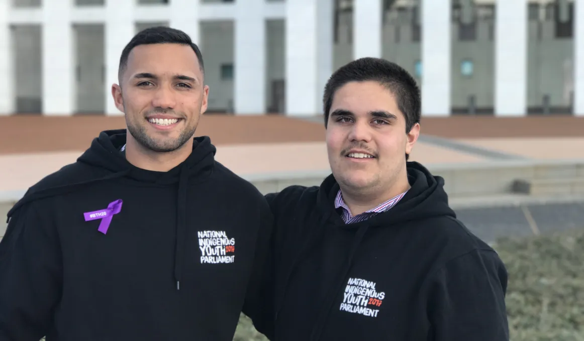 Two young Indigenous men dressed in black hoodies stand arm in arm in front of the white pillars of the Australian Parliament House. The text on their jackets read National Indigenous Youth Parliament.