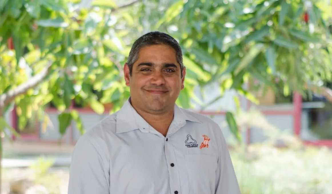 Indigenous man in light coloured shirt stands in front of green tree