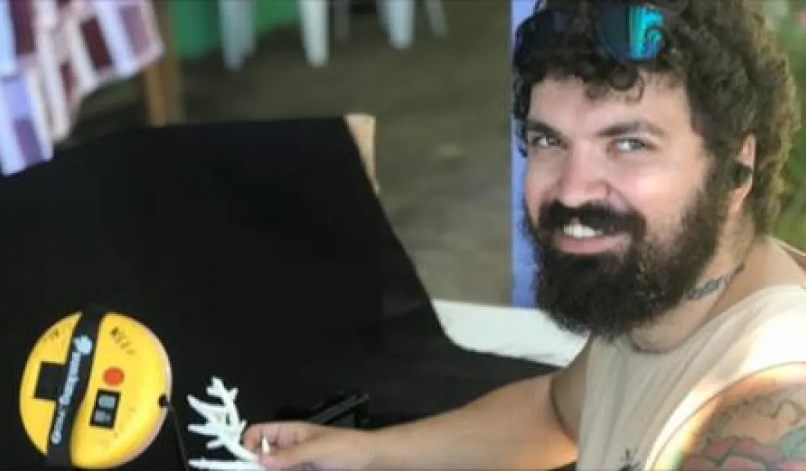 Aboriginal man with curly hair and beard wearing a pale top holds some coral next to an electronic measuring device.