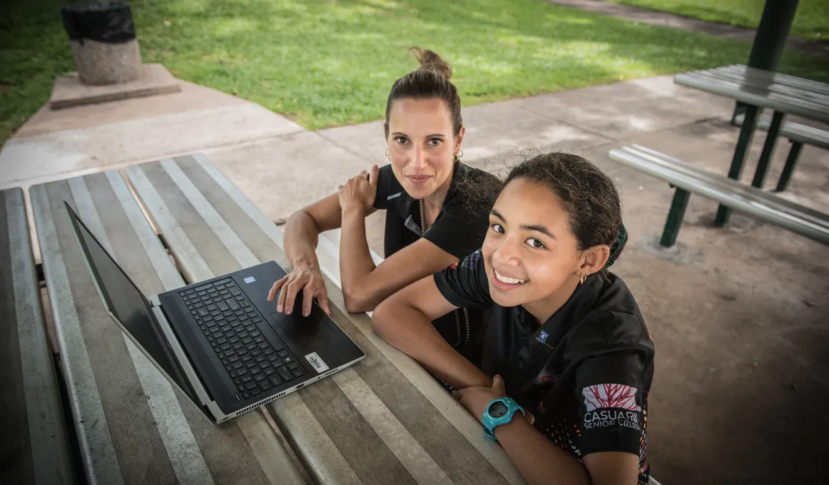 An Indigenous woman and teenaged girl sit at an outside table bench and table. In the background is a bin and green grass.