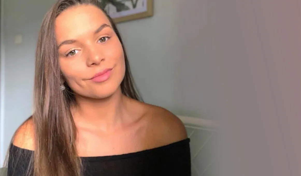 Head and shoulder photo of a young adult woman with long hair and pink lipstick. In the background is a painting hanging on a wall.