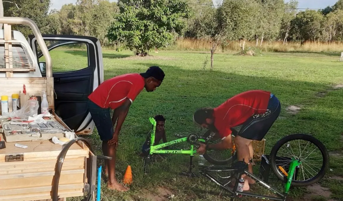A man in red shirt bends over a green pushbike laying on grass. Watching at left is another man in red shirt and behind is a young boy sitting and watching. At left is a truck and in the background is grass and trees.