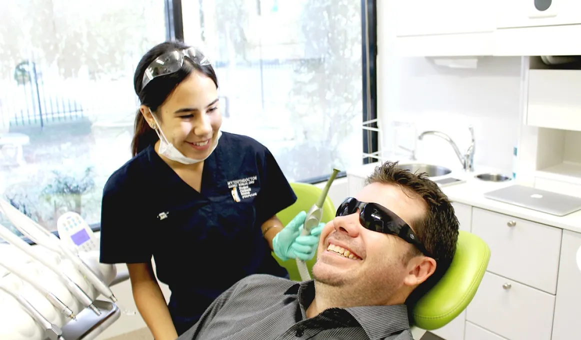 Young Indigenous woman in dark blue uniform stands holding a dentist drill behind a smiling man wearing sunglasses and lying back in a dentist chair.