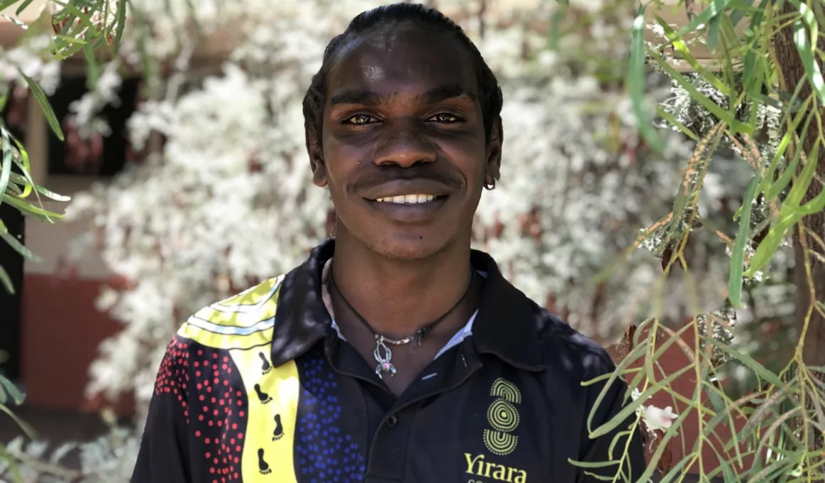 A young man in a polo shirt with traditional designs smiles at the camera. A tree branch with leaves and flowers hangs in front and to his left. In the background is more foliage.