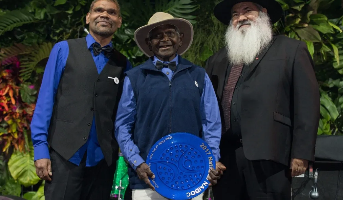 An elderly Aboriginal man holds a large blue plate with a younger Aboriginal man standing to his right and an Aboriginal man with a grey beard standing to his left.