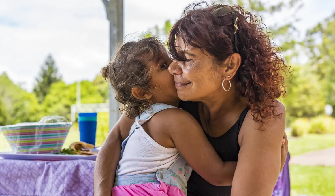 Woman in black top hugs a young girl in pink top. In the background is a table and trees.