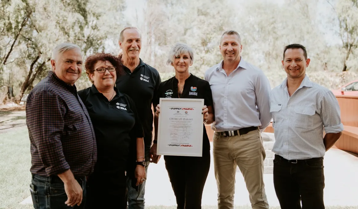 Six people wearing casual clothing stand shoulder to shoulder. In the middle a woman holds up a framed certificate.