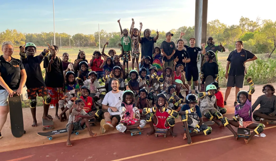 A group of young people and adults stand and sit on a hard surface. Some wear helmets and others hold skateboards. In the background is grass and trees and poles.
