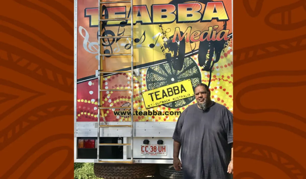 Aboriginal man in grey shirt stands in front of the rear door of a truck. The door is painted with red, yellow and black designs and includes the following words: TEABBA Media, TEABBA N.T. Outback Australia, www.teabba.com