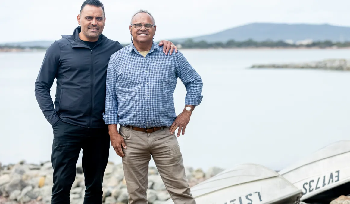 Two men standing in front of a lake. Beside them are two upturned small boats on the rocky shoreline.