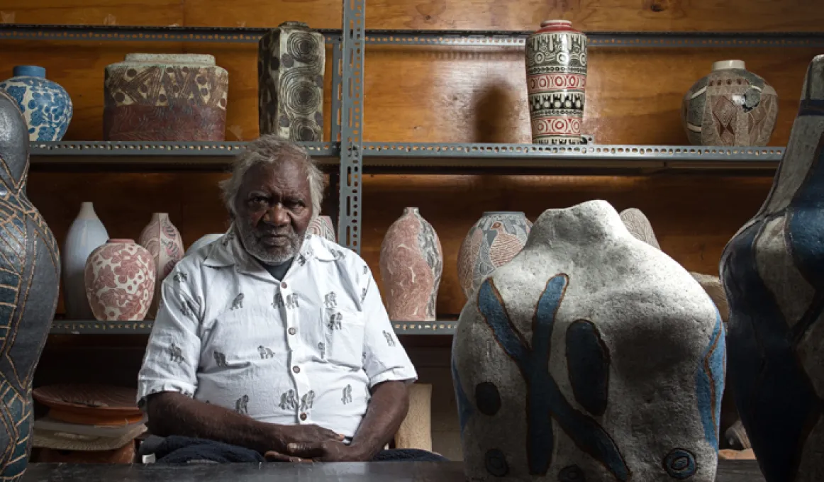 Elderly Indigenous man sits in a room surrounded by large and small pots with Indigenous designs painted or carved on them. Behind him is a wooden wall in front of which is a steel rack upon which are pots.