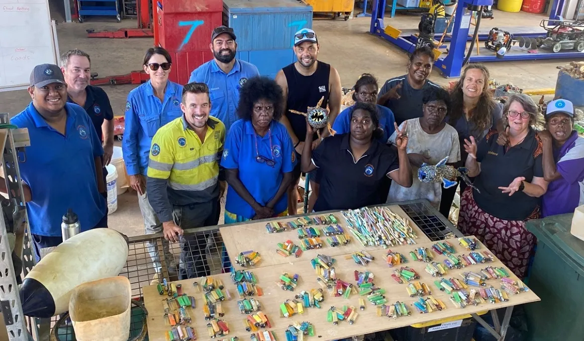 A group of people in a shed stand behind a table covered with cigarette lighters and toothbrushes. Under the table is equipment and in the background are steel racks, cupboards and machinery.