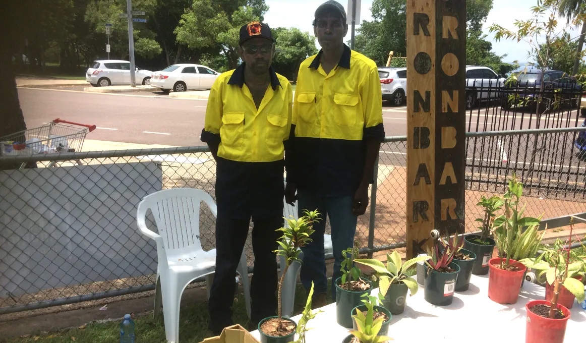Two Aboriginal men standing outside in front of a table with pot plants and fruit. In the background there are trees and cars.