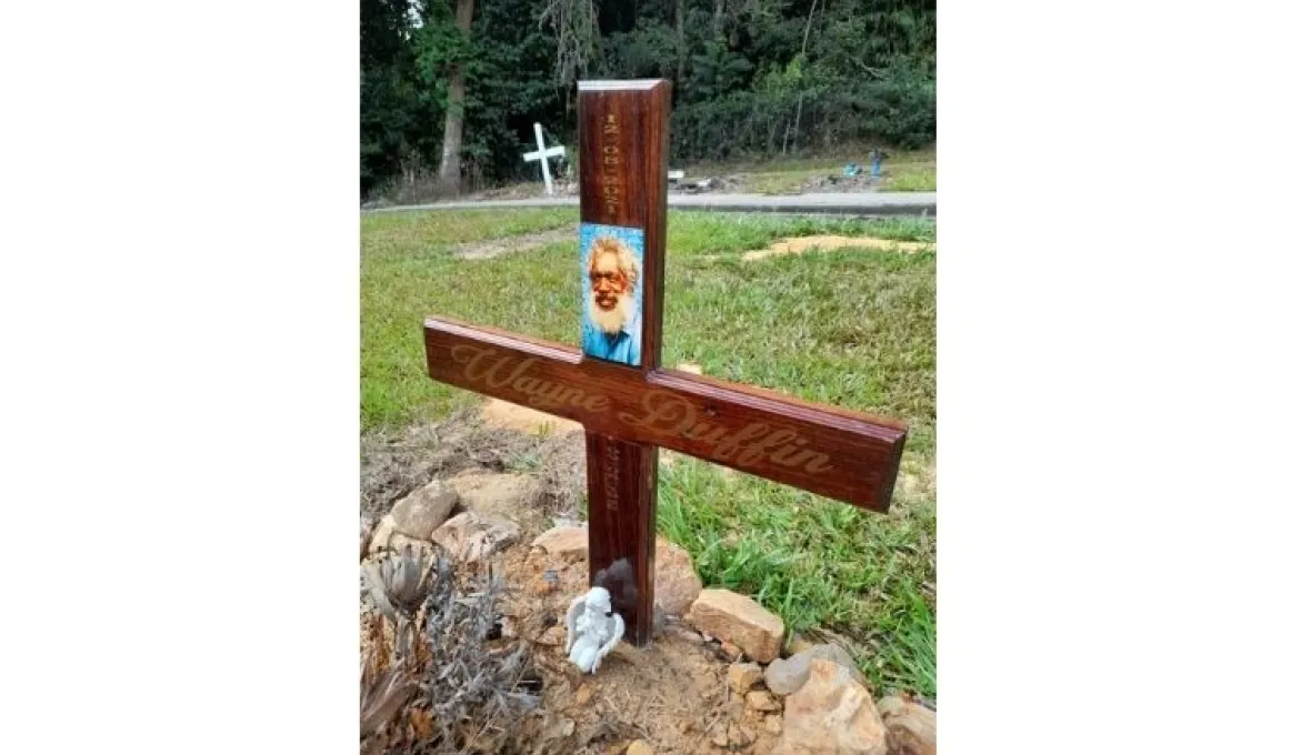 A brown cross sits on a grave. It includes the name and photo of the deceased.