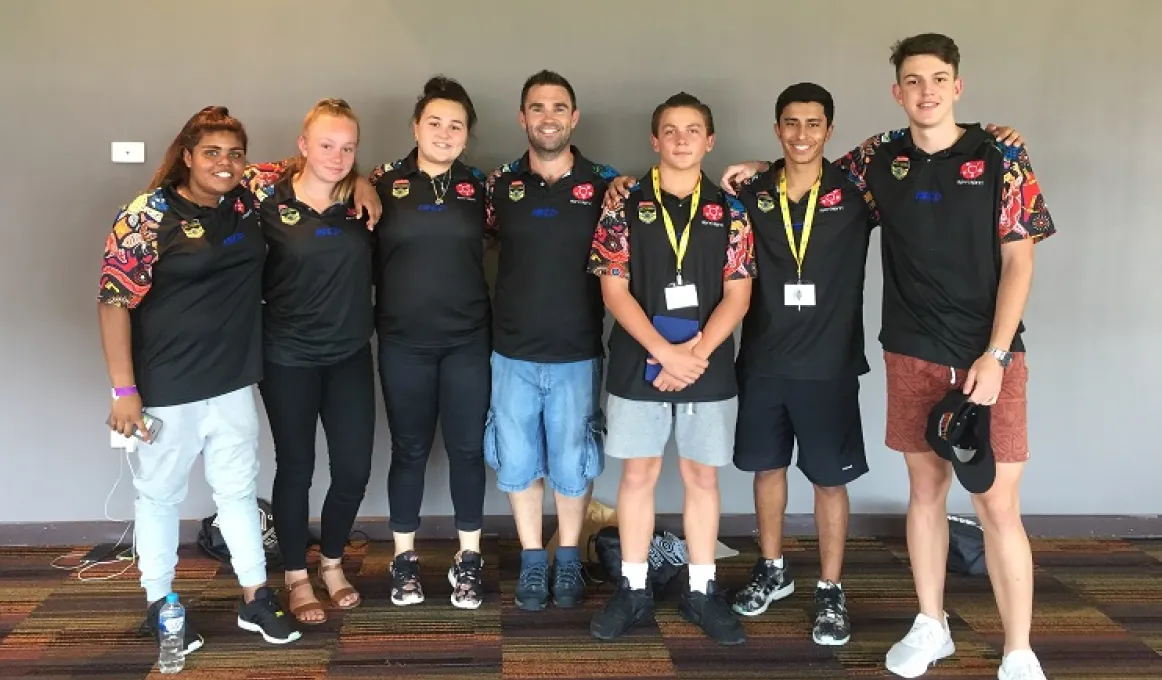Group of six youth and one adult, all Indigenous, wearing black tops and standing arm in arm on a carpeted floor in front of a blank wall.