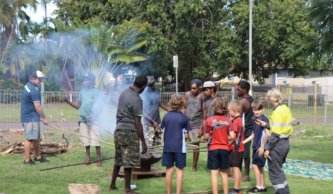 Group of young men stand around a fire placing long sticks over the fire from which smoke arises. In the background are trees, a fence and a light pole.