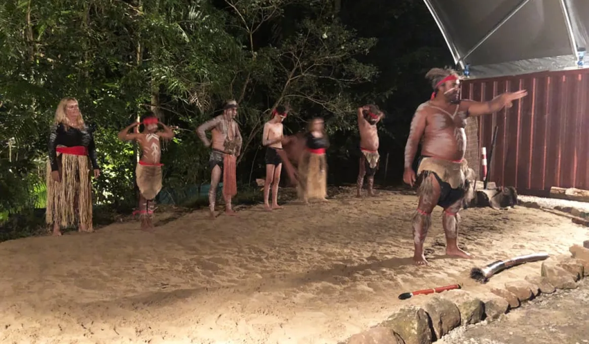 An Indigenous man in traditional wear stands on sand pointing ahead. Behind him are 6 Indigenous people also dressed in traditional wear, 3 of whom are young. In the background is a rainforest and at right, part of a building.