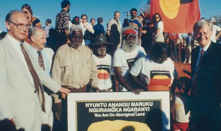 A group of people surround a poster plaque displaying the handing back of the land.