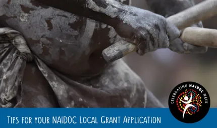 Closeup shot of hands holding sticks which are touching. The hands, arms and belly of an Indigenous person are painted in the traditional way.