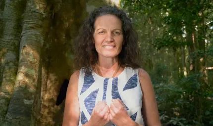 A woman with long dark hair and wearing a blue and white dress stands in a forest. In the background are trees, logs and small shrubs.