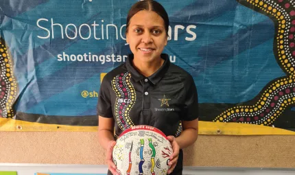 A young woman with her black hair pulled back holds a white ball in her hands. On the ball are traditional Aboriginal designs. In the background is a blue poster with the words Shooting Stars and some more Aboriginal artwork.