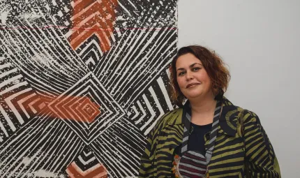 A woman in a green and black striped coat stands next to a mixed media screen print on the wall. It is black, white and ochre in colour with lines and chevrons radiating from a centre made of concentric squares.