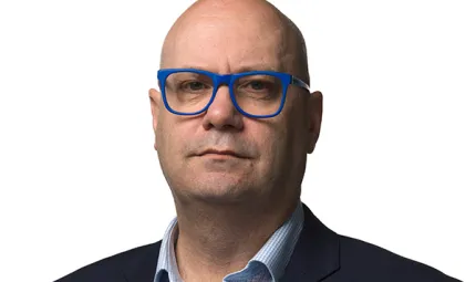 AIATSIS CEO, Craig Ritchie, posing in front of a white background with blue framed glasses, light blue shirt and navy blue blazer.