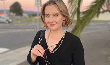 A young woman in a black top holds her glasses in front and smiles at the camera. In the background are tree branches, a car and some signposts.