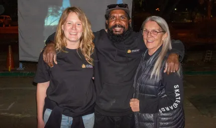 Two women and a man stand shoulder to shoulder. All are dressed in dark clothing. The background is dark except for a pale screen that displays a projected image.