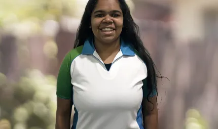 A young adult woman in a green, white and blue top smiles at the camera. She has long black hair. The background has been blurred.