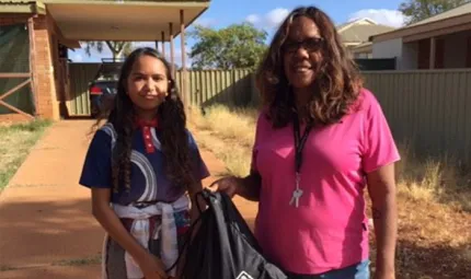 A woman, named Janine Binsiar and a student, Cherice, are standing in a driveway in front of a house and are holding a school and hygiene supply pack.