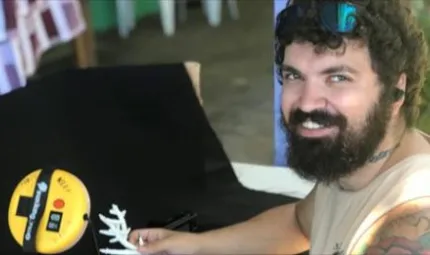 Aboriginal man with curly hair and beard wearing a pale top holds some coral next to an electronic measuring device.
