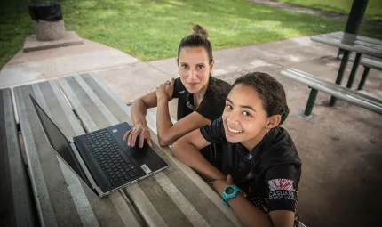An Indigenous woman and teenaged girl sit at an outside table bench and table. In the background is a bin and green grass.