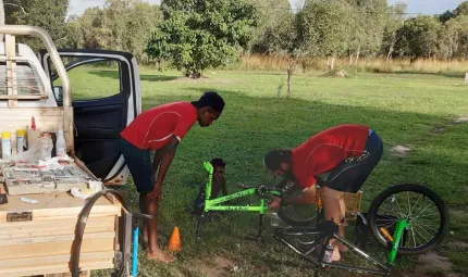 A man in red shirt bends over a green pushbike laying on grass. Watching at left is another man in red shirt and behind is a young boy sitting and watching. At left is a truck and in the background is grass and trees.